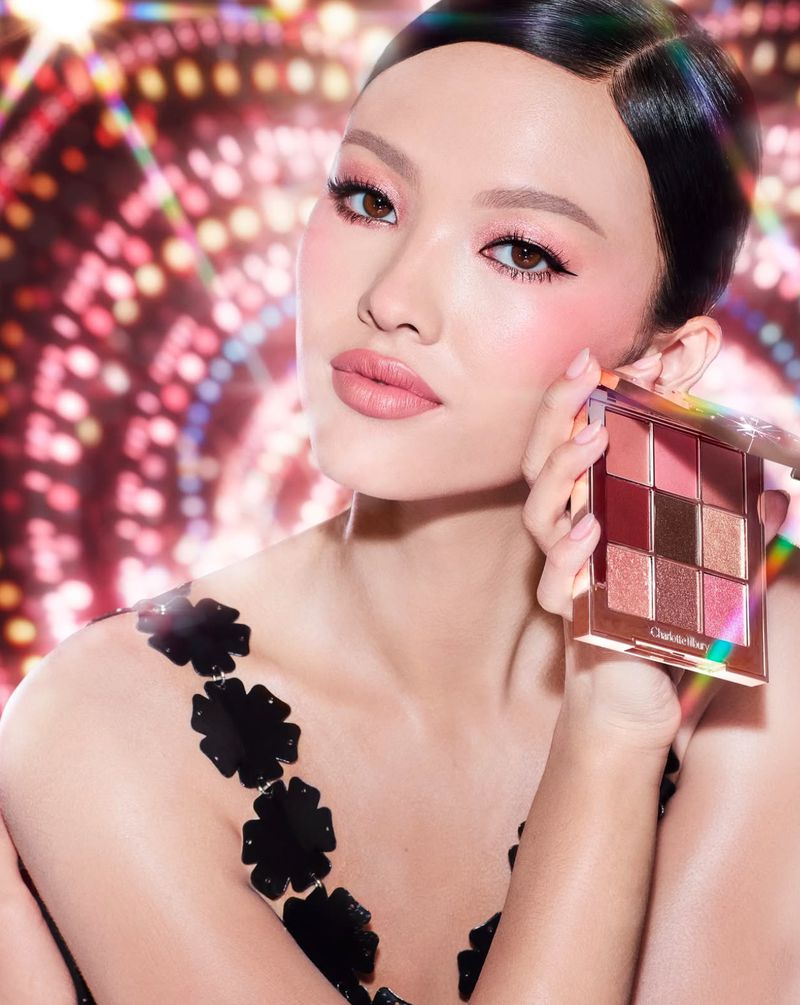 Close-up of a beautiful Asian woman holding a makeup palette, showcasing pink eyeshadow and cheek color, set against a bokeh background.