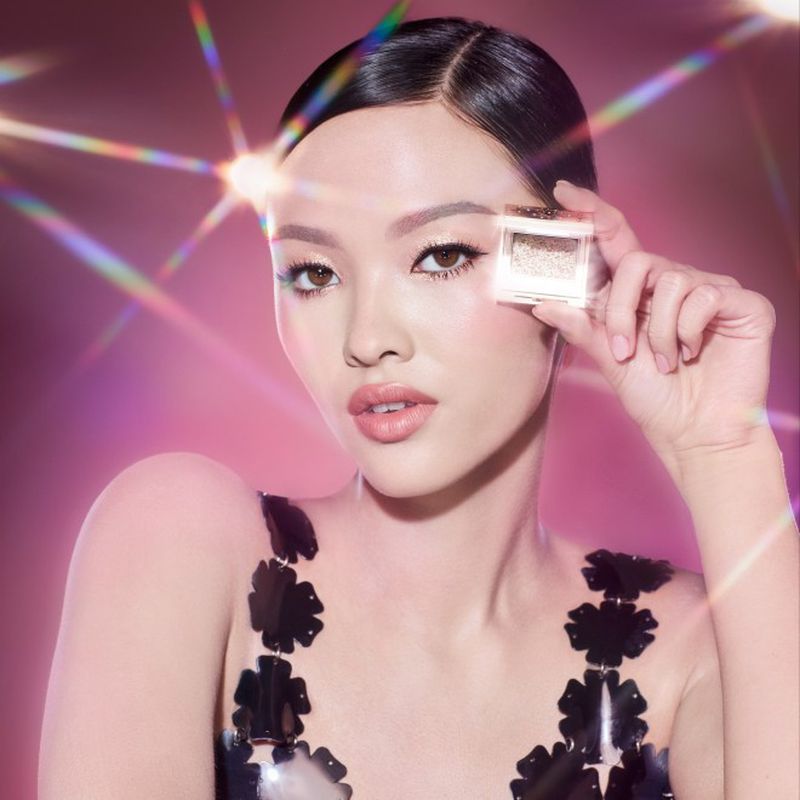 Glamorous portrait of a woman holding shimmering eyeshadow in a compact, showcasing makeup and beauty products against a pink bokeh background.