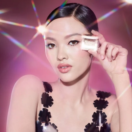 Glamorous portrait of a woman holding shimmering eyeshadow in a compact, showcasing makeup and beauty products against a pink bokeh background.