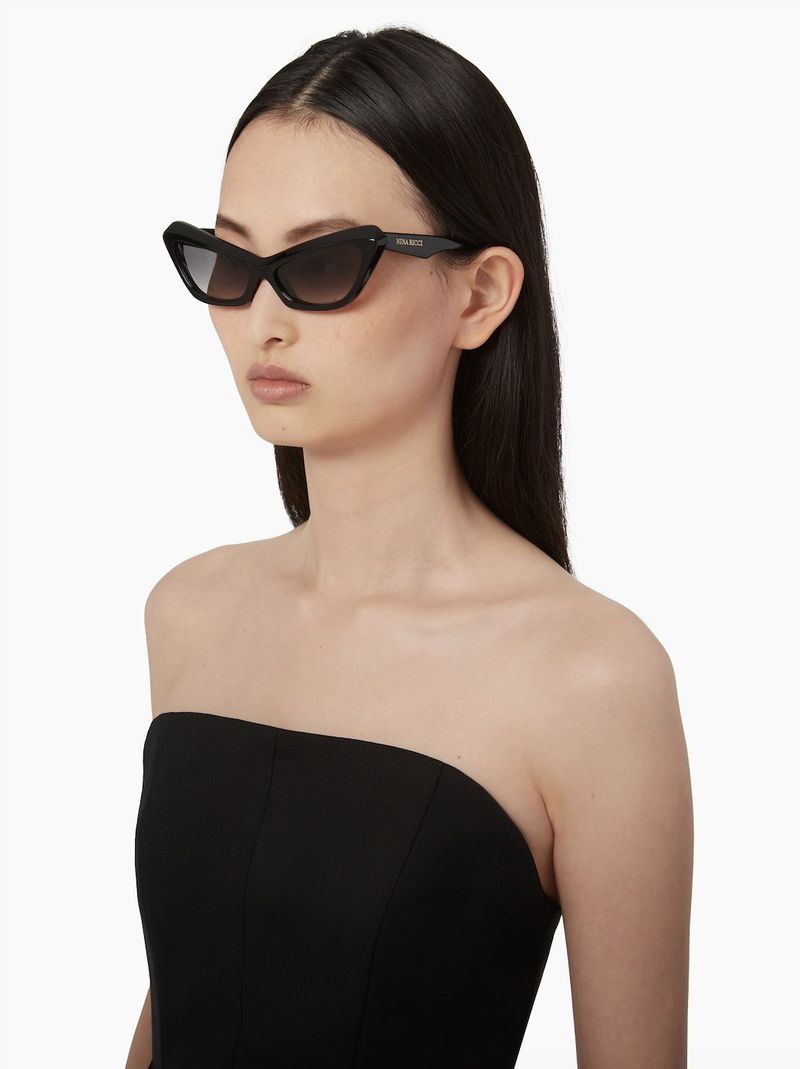 Woman with dark hair wearing black strapless top and black cat eye sunglasses against a white background in a studio shot.