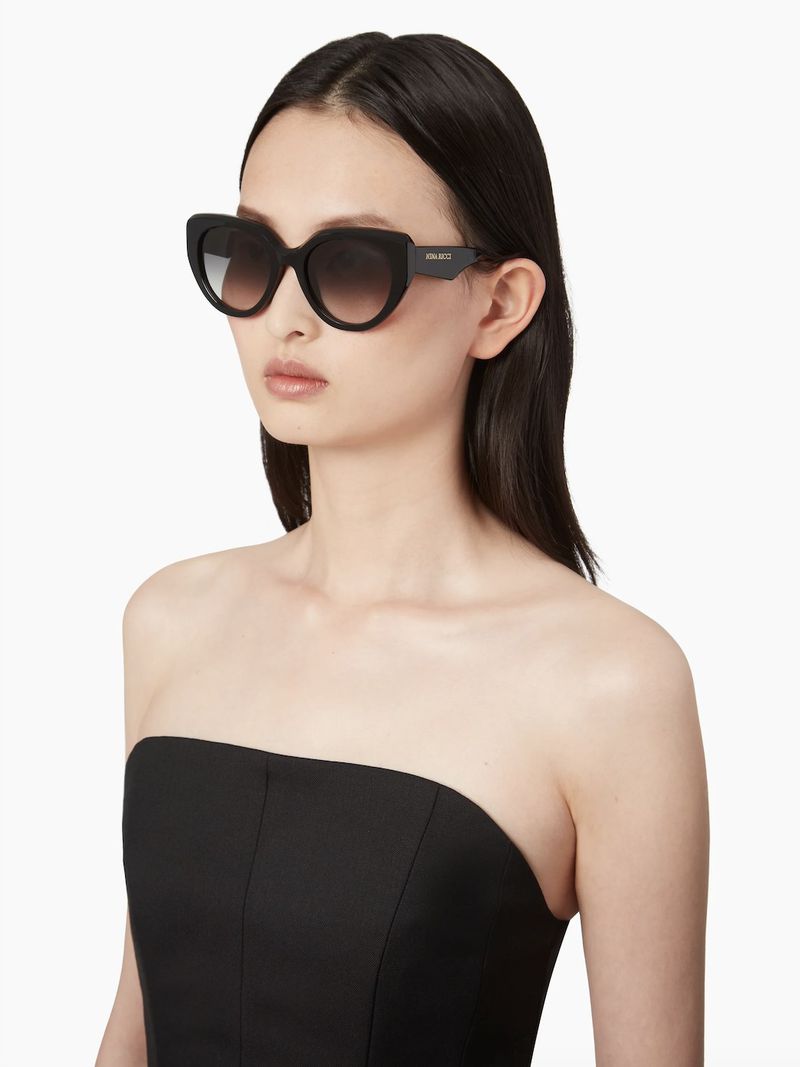 A woman with dark hair wearing black sunglasses and a strapless black top poses against a white background, showcasing a fashion or beauty aesthetic.