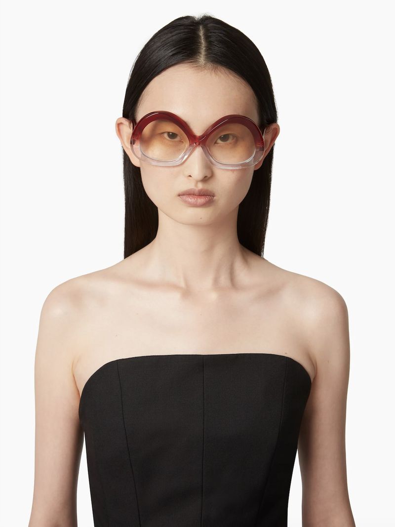 Asian woman poses in a strapless top wearing oversized brown-tinted sunglasses against a white backdrop in a studio-style setting.