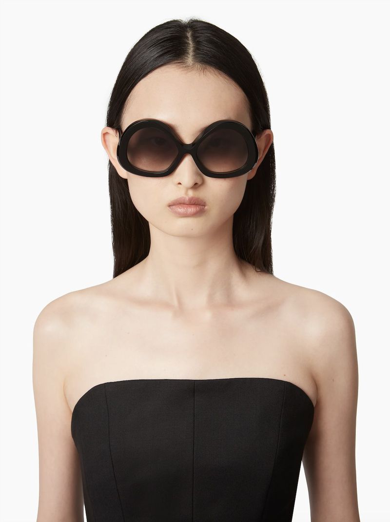 Headshot of a woman wearing black sunglasses and a black strapless top against a white background in a portrait orientation.