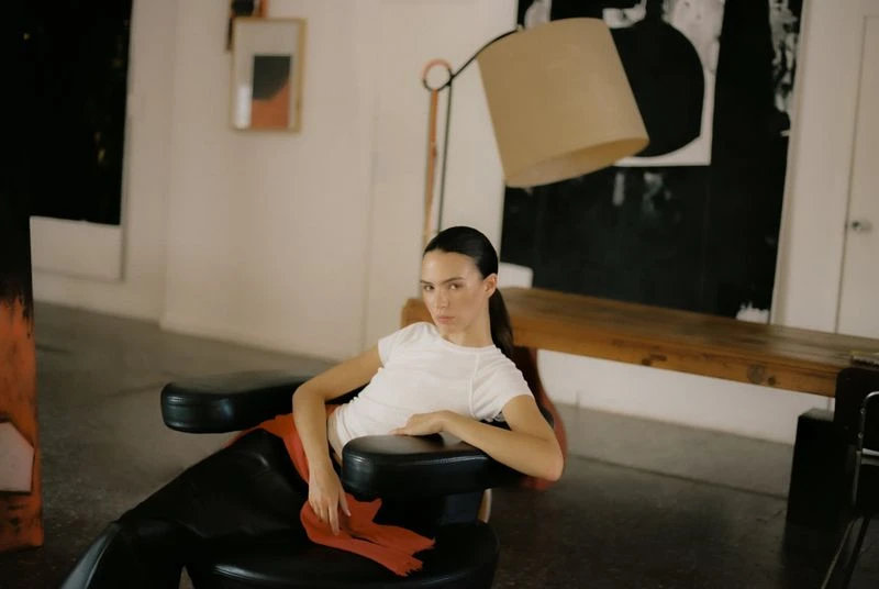 Woman poses in a black chair in a brightly lit studio with an abstract painting and floor lamp in the background. Fashion shot.