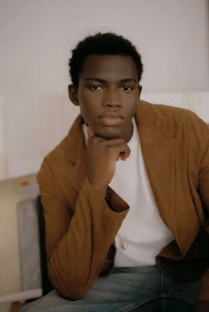 Portrait of an adult African American man with brown hair, wearing a brown jacket and white shirt, seated indoors with hand to face.