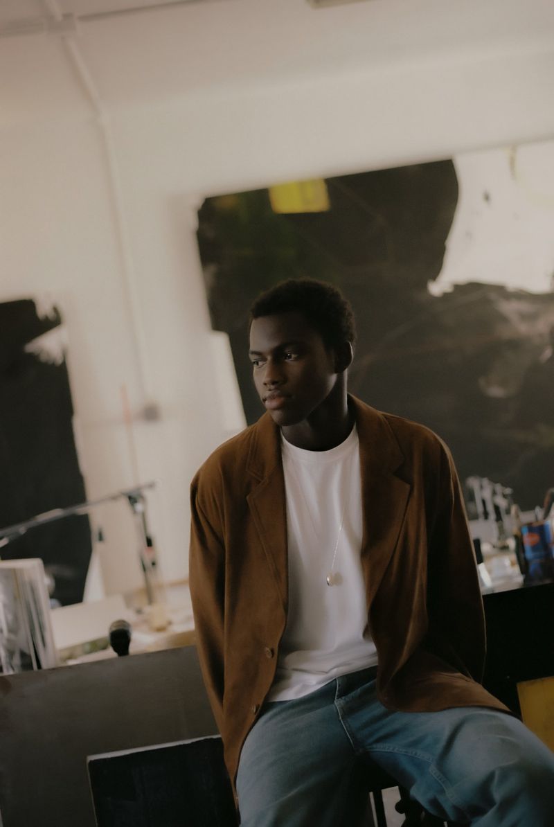 Young man in a brown jacket sits in a room with art; he wears jeans and a white shirt, looking off to the side, casual and relaxed.