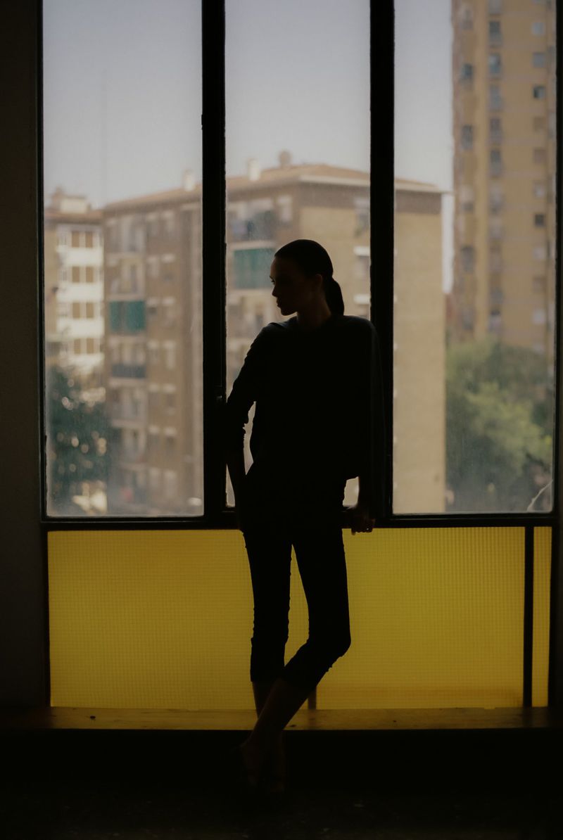 Silhouette of an adult woman standing by a window with a cityscape view in the background, featuring natural light and dark tones.