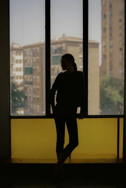 Silhouette of an adult woman standing by a window with a cityscape view in the background, featuring natural light and dark tones.