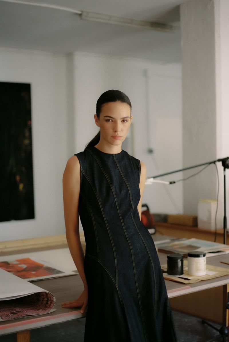 A young woman in a black dress poses in an artist's studio with painting and art supplies in a naturally lit, indoor setting.