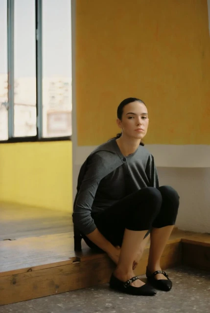 Woman sits in gray top and black leggings near a bright window, illuminated by natural light in an indoor environment.