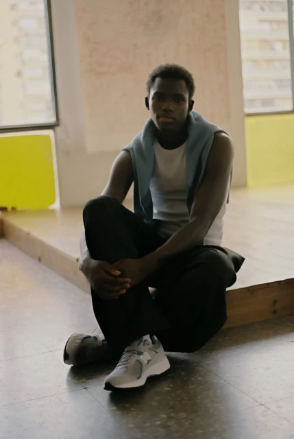 Young adult man sits indoors wearing casual clothes and sports sneakers, looking at the camera with a neutral facial expression.
