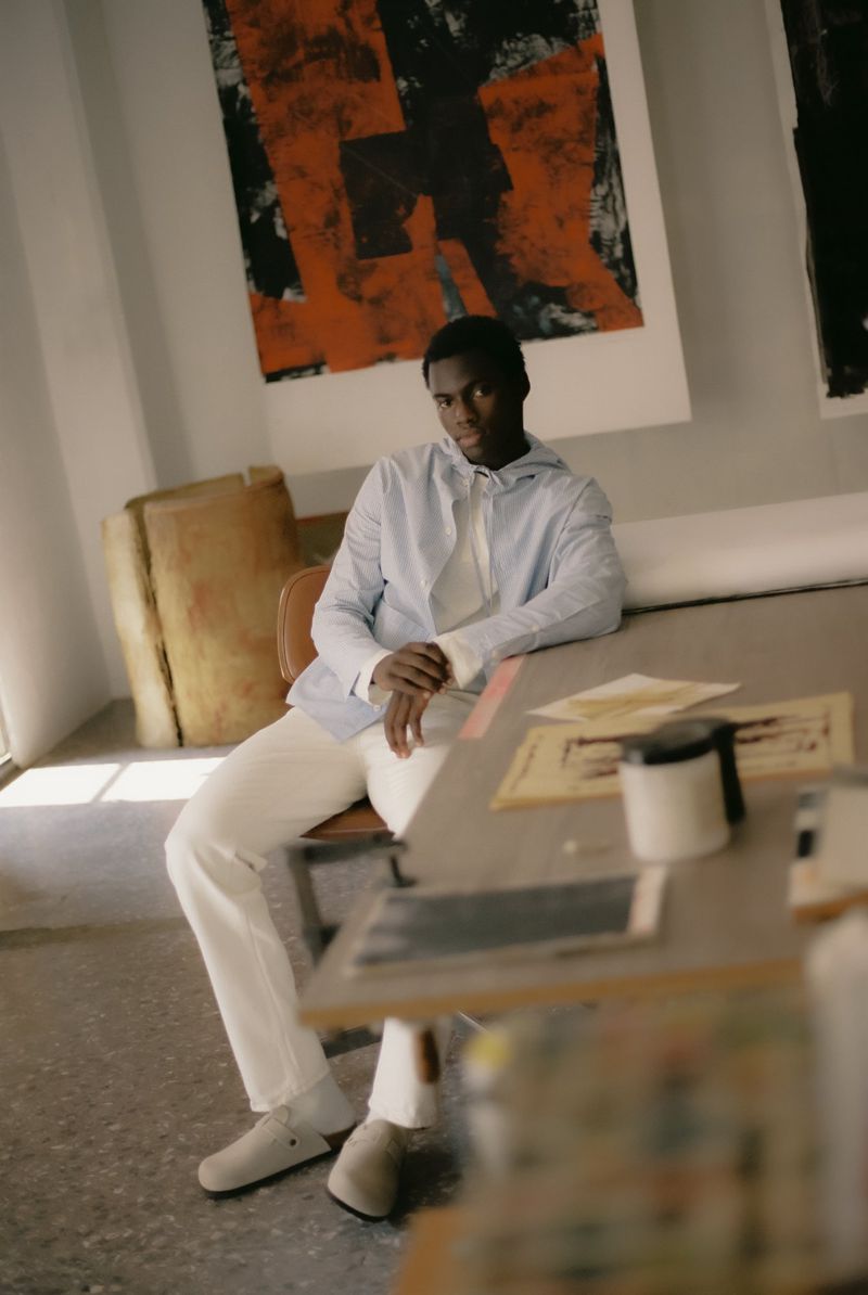 Adult male sitting at a table in a light blue shirt and white pants in an indoor setting with abstract art on the walls.