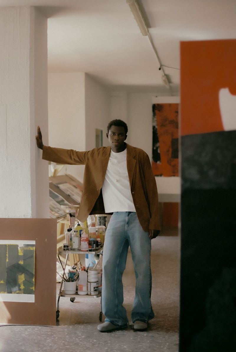 A casually dressed Black male model poses in an artist's studio, leaning against a white wall next to an abstract painting on a cart.