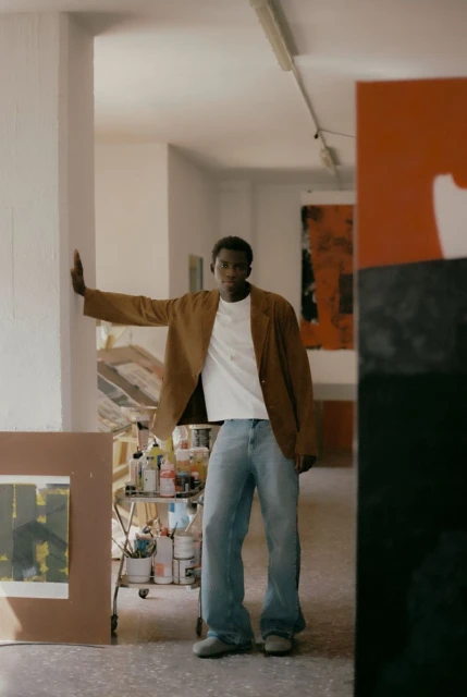 A casually dressed Black male model poses in an artist's studio, leaning against a white wall next to an abstract painting on a cart.