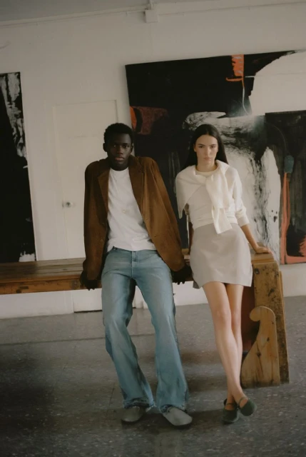 Two fashion models in a light-filled photo studio. A Black man in jeans and a brown jacket poses with a White woman in a skirt and blouse.