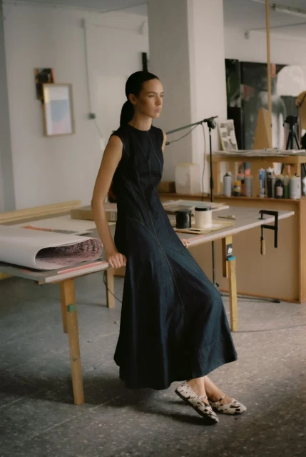 A slender woman in a dark sleeveless dress leans against a light wooden table in a brightly lit photo studio with painting supplies.