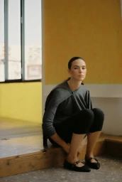 Woman in grey top and black trousers seated on wooden flooring against a yellow and white interior wall.