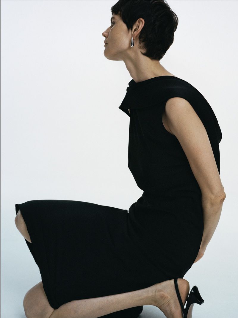 Woman kneeling in a black dress against a white background. The model is in a simple, elegant pose and style.