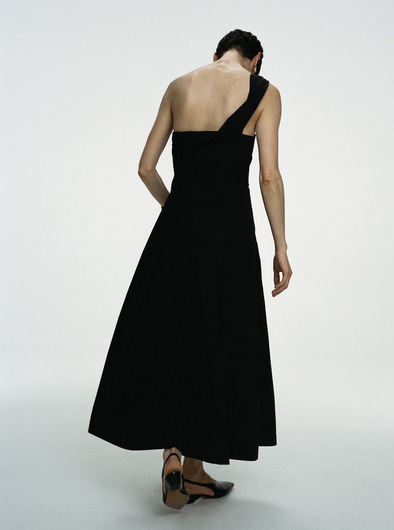 Model wearing a black one-shoulder dress seen from behind against a plain white studio background.