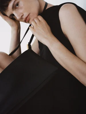 Model in a black dress holding a black leather handbag against a plain white background