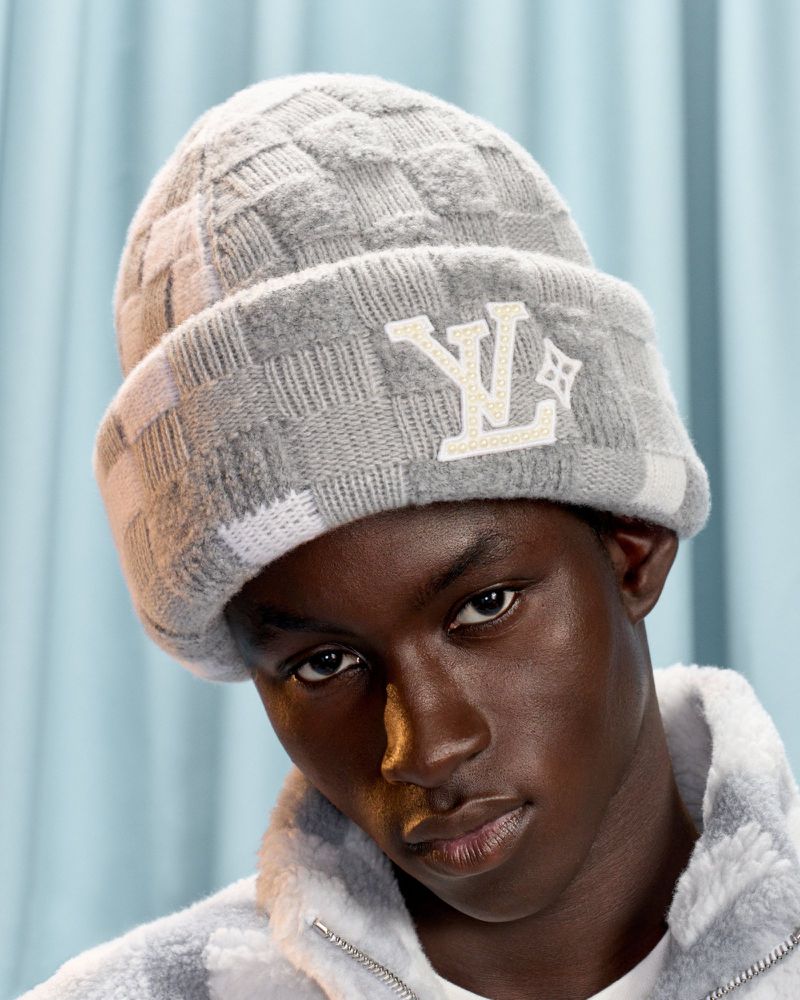 Close-up portrait of a young adult male with dark skin wearing a grey checkered beanie with the LV logo against a blue background.