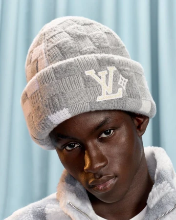 Close-up portrait of a young adult male with dark skin wearing a grey checkered beanie with the LV logo against a blue background.