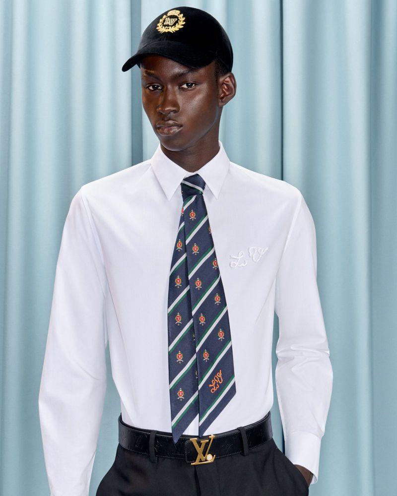 Portrait of a Black male model wearing a white dress shirt, patterned tie, black pants, and a cap against a light blue backdrop.