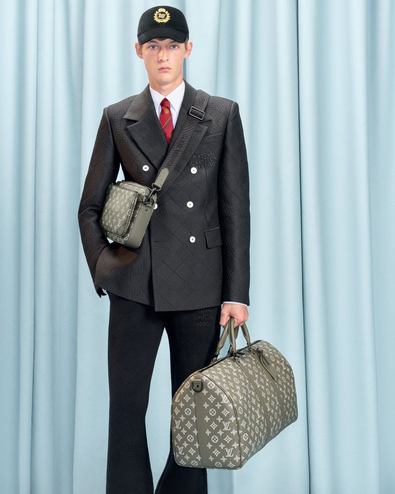 A young male model poses indoors against a light blue backdrop wearing a black suit, baseball cap, and holding two patterned duffel bags.