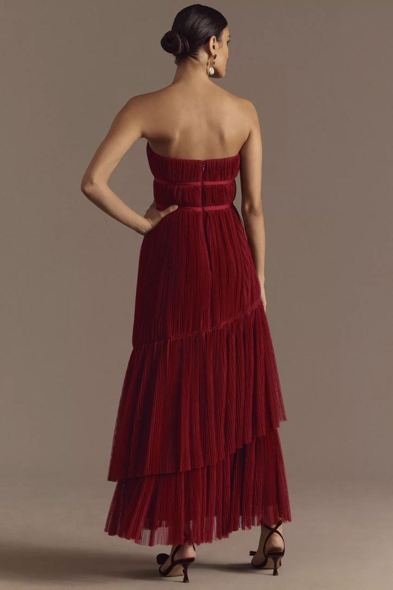 Back view of a woman in a strapless, tiered, deep red dress and heels, posing in front of a neutral studio backdrop.