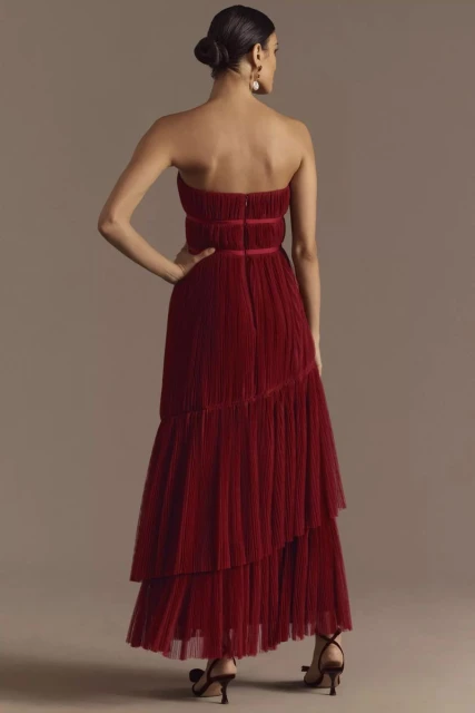 Back view of a woman in a strapless, tiered, deep red dress and heels, posing in front of a neutral studio backdrop.