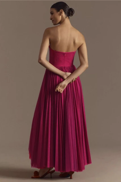 An adult woman stands with her back to the camera, wearing a floor-length fuchsia dress with a strapless bodice in a studio setting.