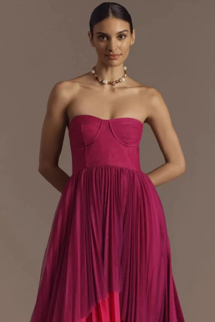 Brunette woman posing in a fuchsia strapless dress with a tulle skirt and a pearl necklace in front of a neutral backdrop.