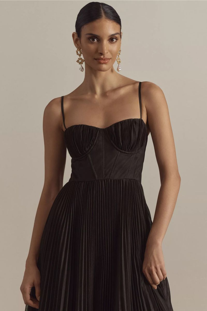 Woman in elegant black dress with pleated skirt and strappy bodice, accessorized with dangling pearl earrings, posing in a studio.