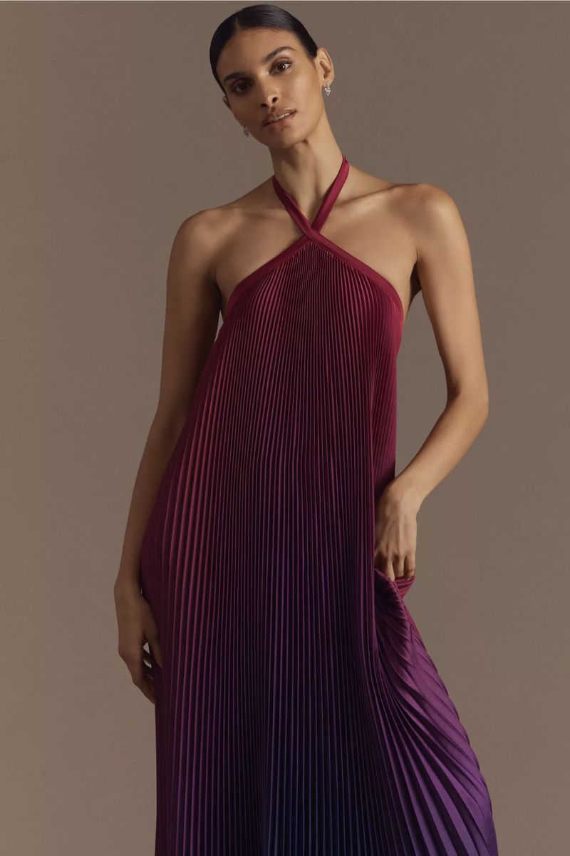 Adult woman wearing a sleeveless, floor-length pleated dress that transitions from burgundy to purple stands in a neutral studio.