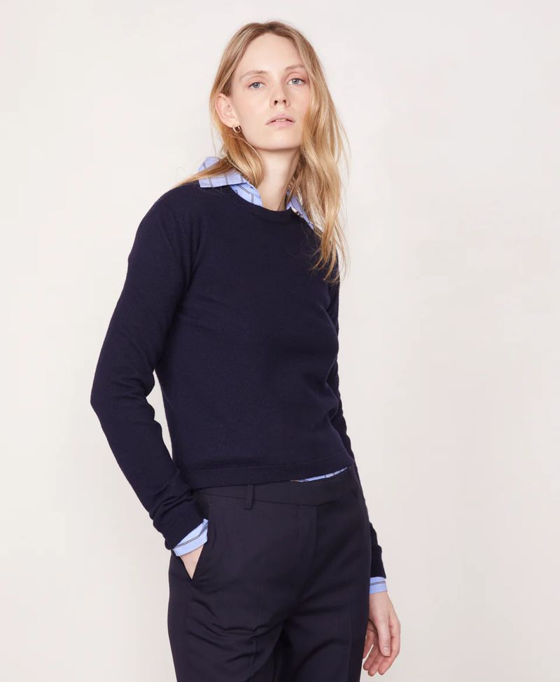 Fashion shot of a blond woman wearing a navy sweater and pants, standing in front of a white background in a studio setting.