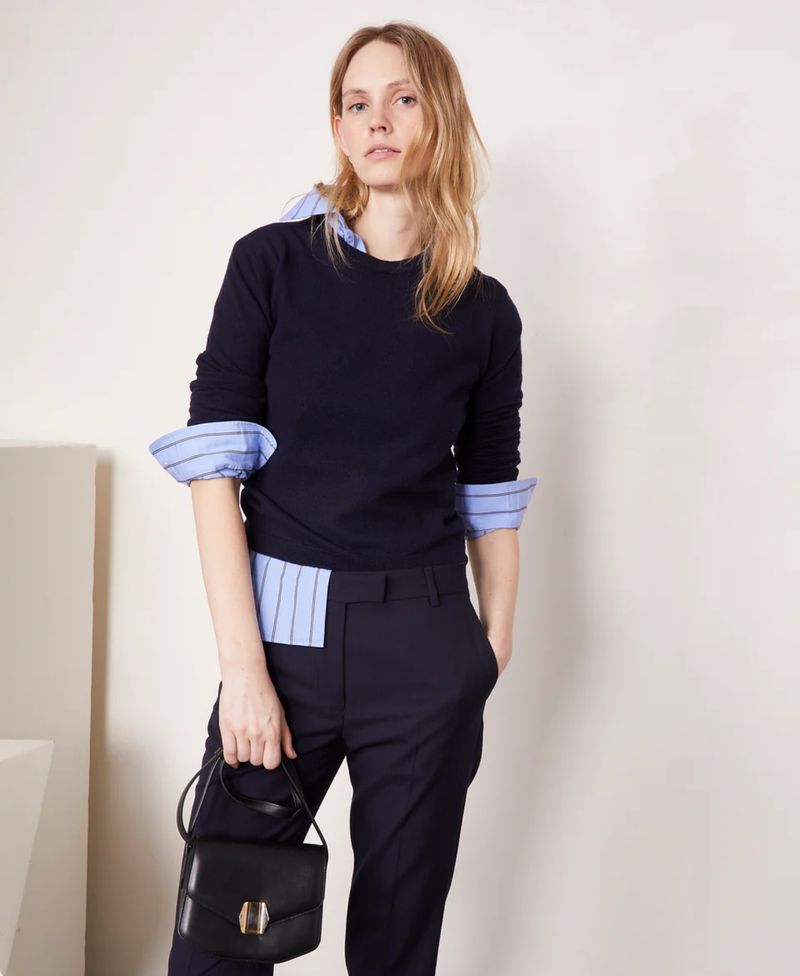Blonde woman in a navy sweater and dress pants holds a small black purse against a neutral studio backdrop, showcasing fashion.