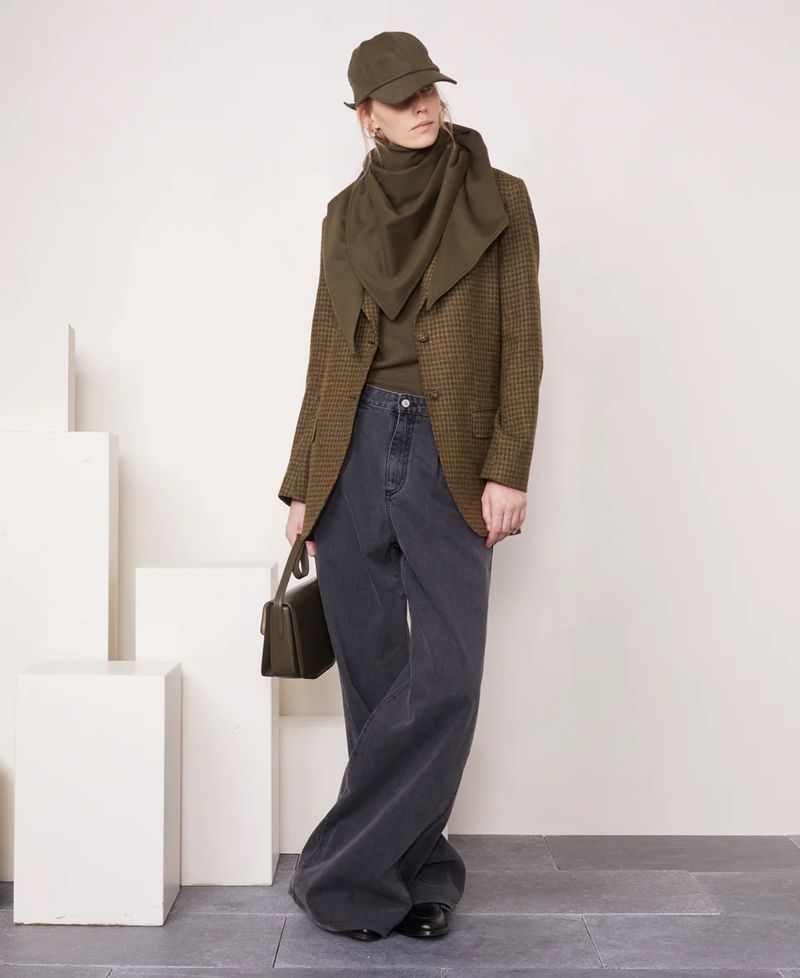 Woman in tweed blazer, grey wide-leg jeans, and green accessories poses in a studio with white blocks and grey flooring.