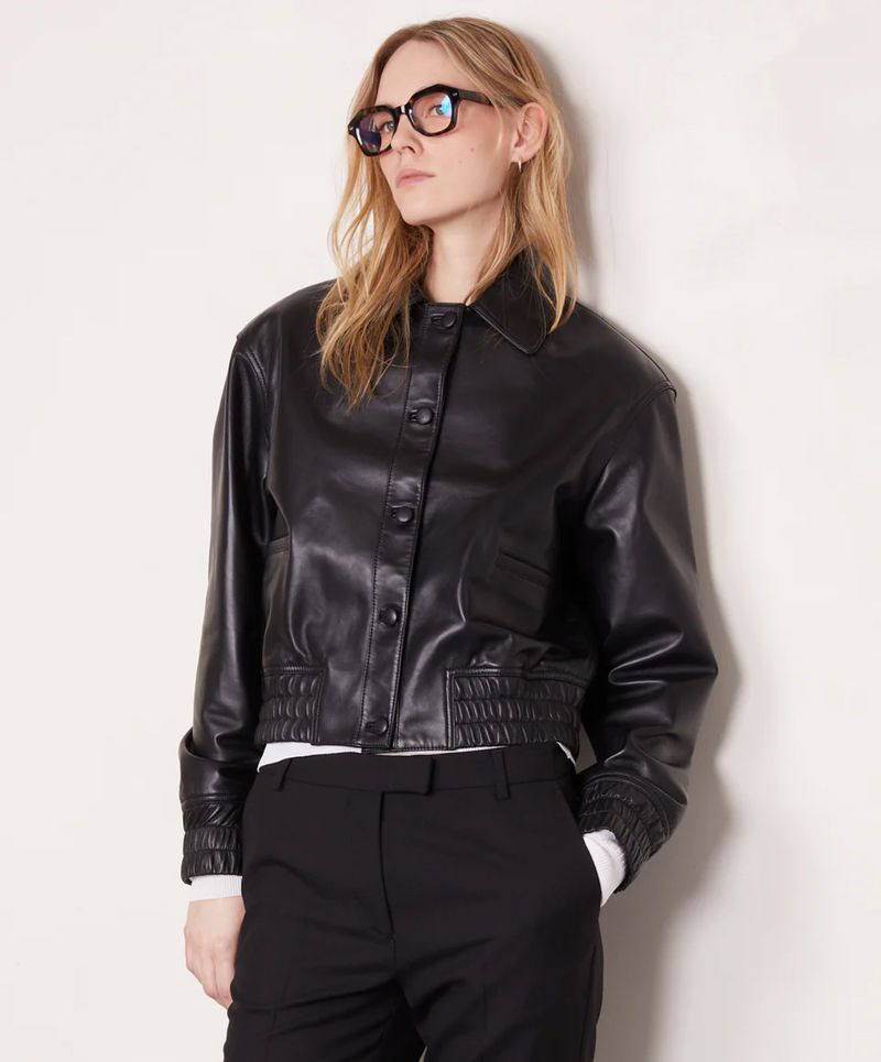 A blonde adult woman in black framed glasses poses in a studio, wearing a leather jacket and black pants. The image uses soft lighting.