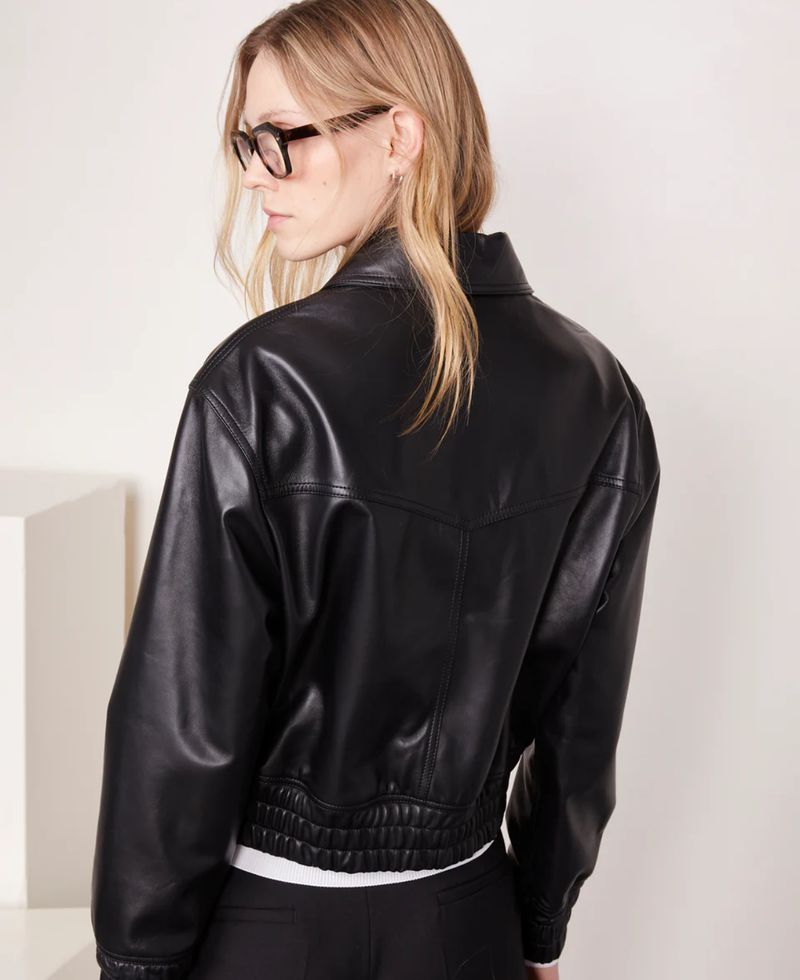 Model in a black leather bomber jacket, wearing eyeglasses, posing in front of a white wall in a studio. Fashion shot.