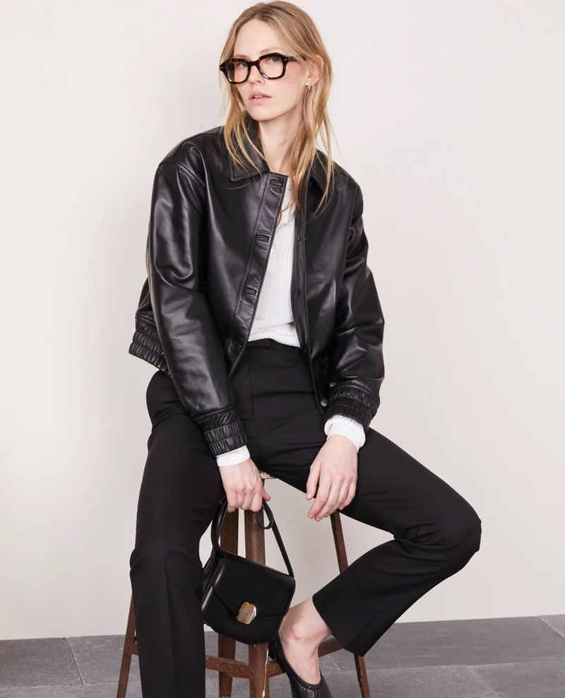 Adult female fashion model in a black leather jacket poses on a stool with a black purse against a neutral background in a studio shot.