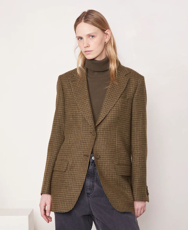 A fair-skinned woman with long blonde hair poses indoors, wearing a brown-green tweed blazer over a dark green turtleneck sweater.