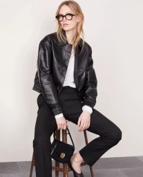 Model wearing black leather jacket, black trousers, and dark glasses sitting on wooden stool holding handbag