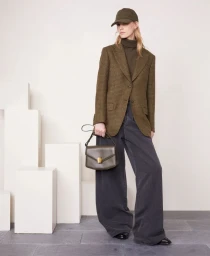 Model wearing an olive tweed blazer, turtleneck, cap, and holding a handbag against a minimalist architectural background.