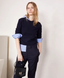 Model in navy sweater and dark trousers holding a black handbag against a plain white studio background.