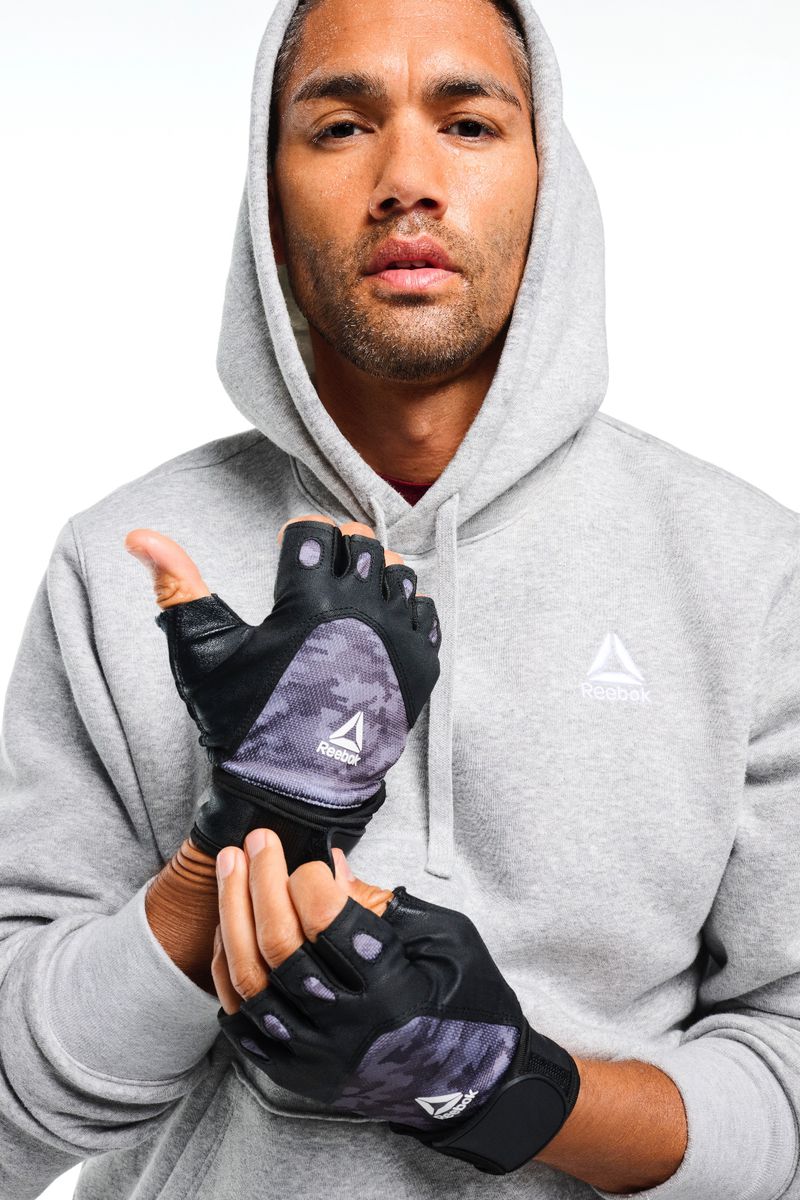 Man wearing fingerless Reebok workout gloves and grey hooded sweatshirt poses in a studio setting with a white background.