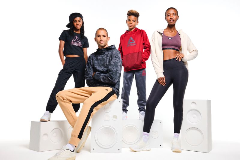 Four diverse people wearing Reebok apparel posing in a studio standing and sitting on speaker props. Reebok logo visible on clothing.