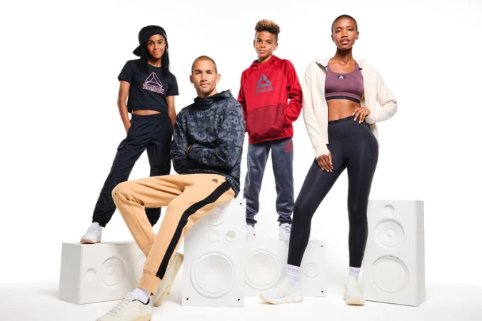 Four diverse people wearing Reebok apparel posing in a studio standing and sitting on speaker props. Reebok logo visible on clothing.