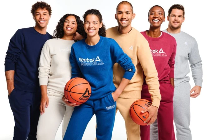 Group of six diverse adult models in matching Reebok tracksuits smile in a brightly lit photo studio, holding basketballs, showcasing sportswear.