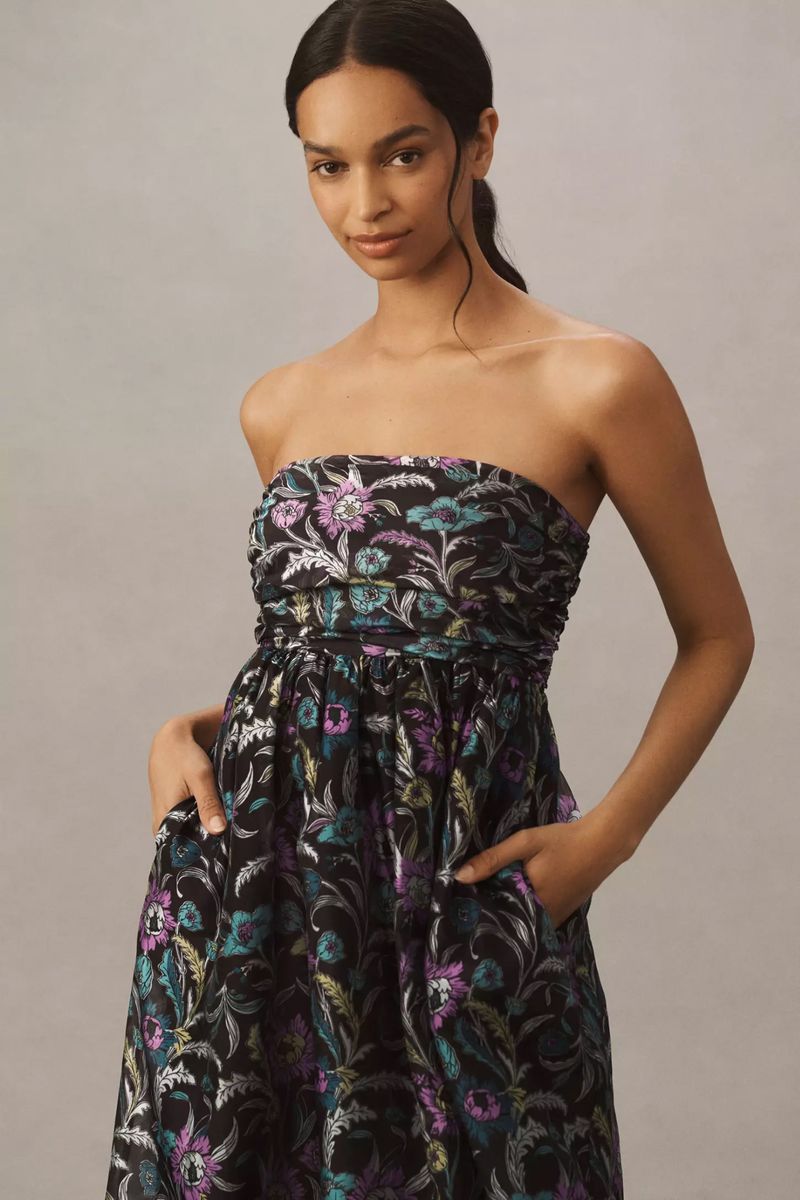 A woman in a strapless floral midi dress with a black background stands indoors with her hands in the pockets of the dress.