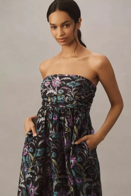 A woman in a strapless floral midi dress with a black background stands indoors with her hands in the pockets of the dress.